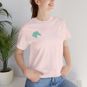 Woman wearing light pink t shirt with minimalist aqua horse head print on left chest standing indoors by a plant