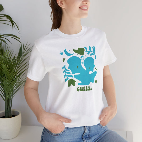White zodiac t shirt featuring a retro blue Gemini twin graphic with green leaves and stars, worn by a smiling woman with hands in pockets.